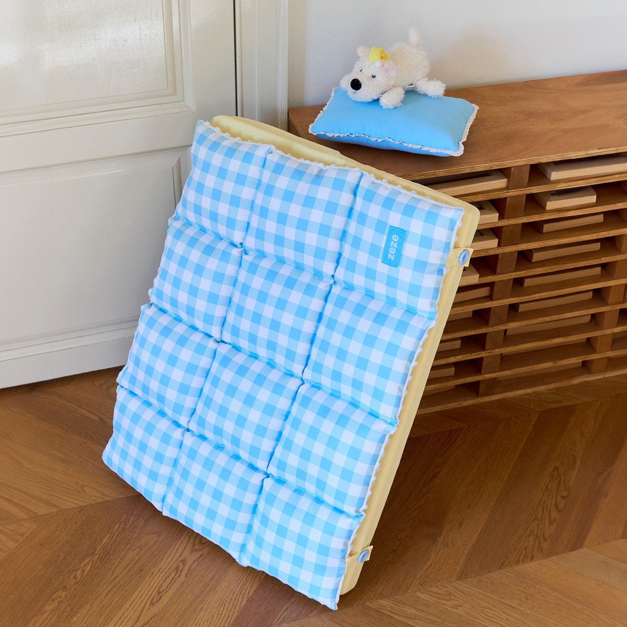 Gingham nap mat standing against a cabinet with pillow and plush puppy