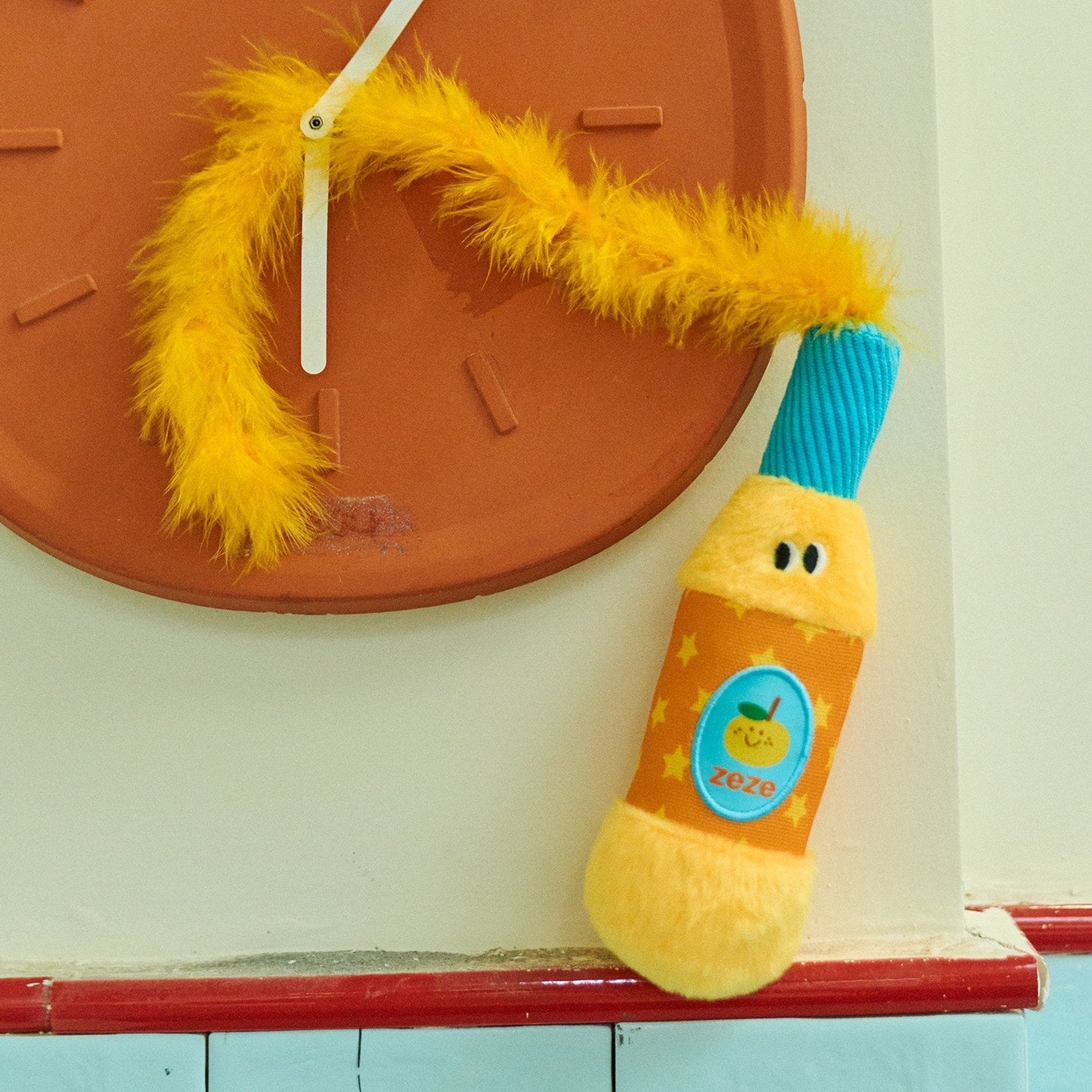 Orange Popper cat toy with its feather tail over a wall clock